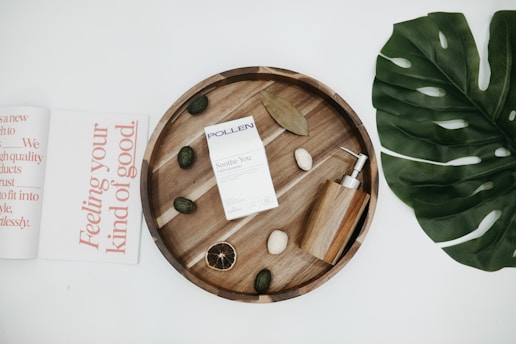 A flat lay featuring a circular wooden tray with various items including a box labeled 'POLLEN Soothe You CBD Gummies', an elegant wooden soap dispenser, and some decorative stones and dried elements. To the left, there is an open magazine with large text in coral font, and to the right, a large green leaf.
