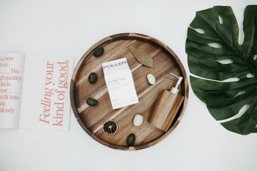 A flat lay featuring a circular wooden tray with various items including a box labeled 'POLLEN Soothe You CBD Gummies', an elegant wooden soap dispenser, and some decorative stones and dried elements. To the left, there is an open magazine with large text in coral font, and to the right, a large green leaf.