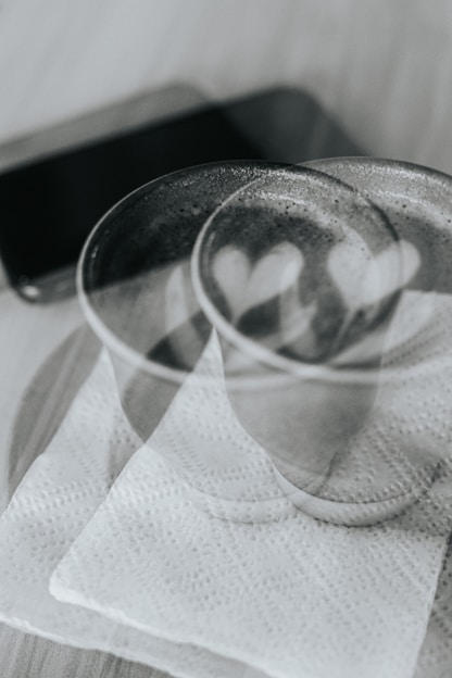 An artistic double exposure photograph featuring two cups placed on a napkin with two heart shapes visible inside the cups. The background shows a blurred mobile phone lying on the table.