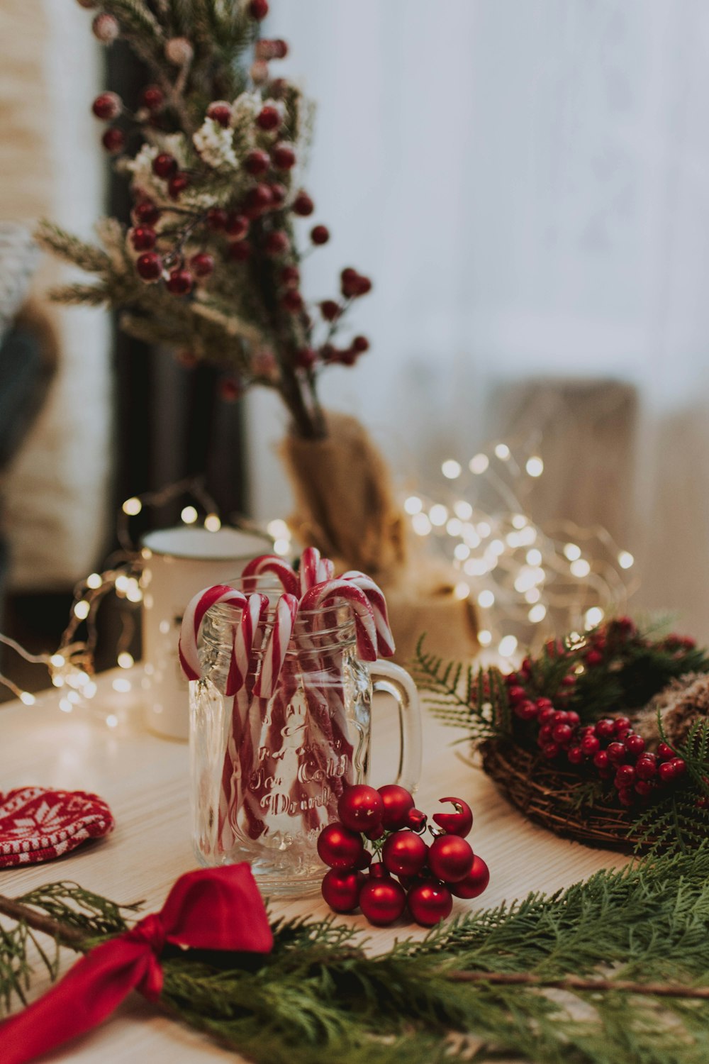 candy cane in clear glass jar near red baubles, lighted string lights, and white mug