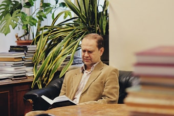 A person is sitting in a comfortable chair reading a book, surrounded by a pleasant atmosphere with indoor plants and stacks of books in the background.