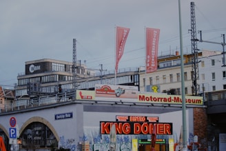 An urban scene featuring a building with various signage, including 'Motorrad-Museum' and 'Bistro'. The area is surrounded by railway infrastructure with visible tracks and overhead wires. The facade is marked with graffiti, and there are flags displaying the museum's name. In the background, there are modern office buildings.