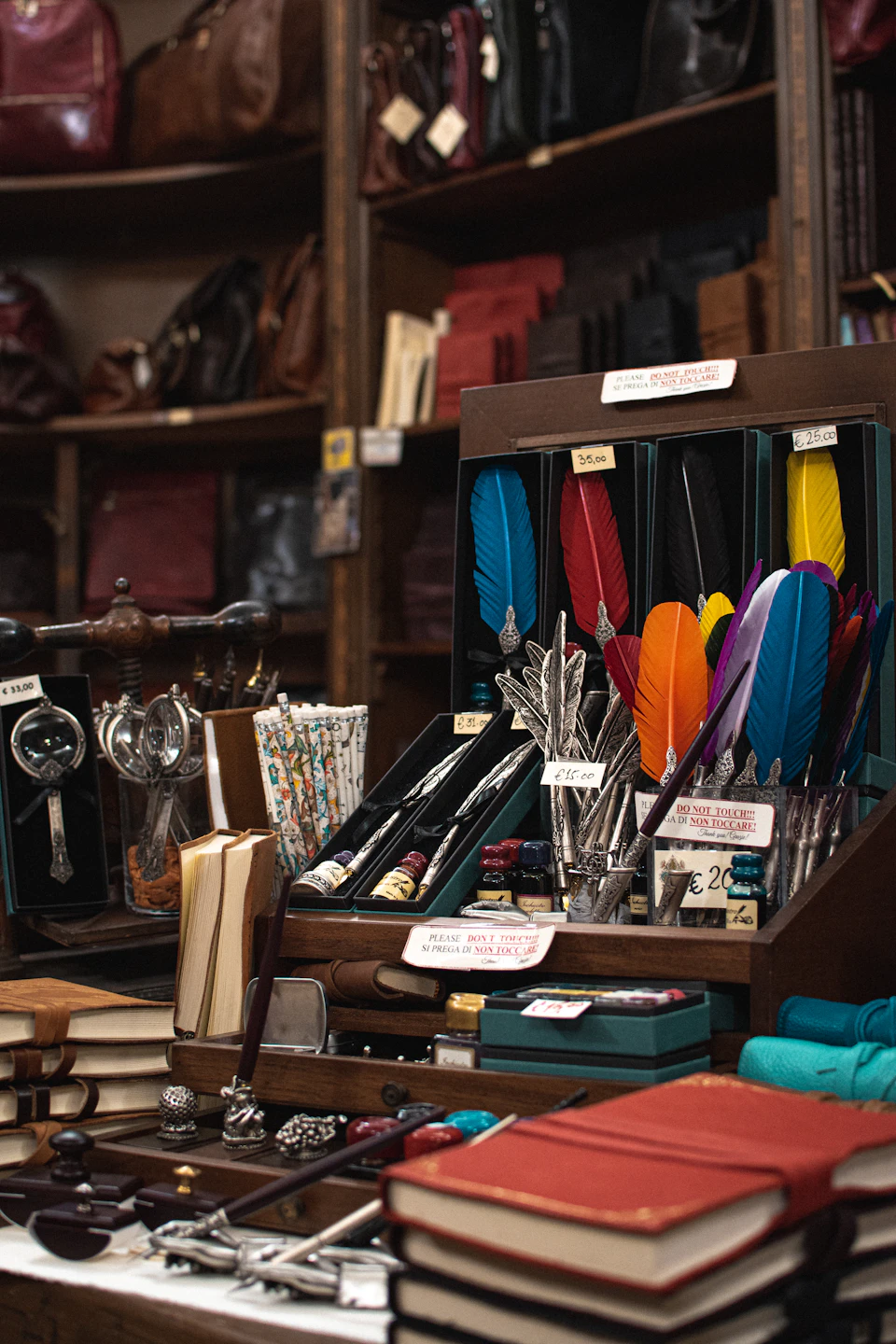 The image depicts a wooden display case filled with an array of colorful feathers and stationery items, including pens, notebooks, and books. The feathers are arranged in various colors such as blue, orange, green, and purple, creating a vibrant and eye-catching presentation. The stationery items include pens, pencils, and markers, all neatly organized within the display case. In the background, there is a shelf filled with more books, adding to the overall sense of a well-stocked store or shop.