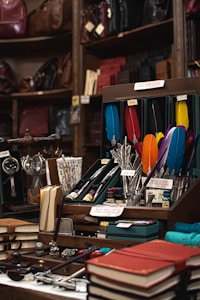 A vintage display of stationery items, including colorful feather quill pens, ink bottles, and notebooks, arranged neatly on a wooden counter. The background features shelves filled with leather bags and bound books, creating a classic and old-fashioned ambiance.
