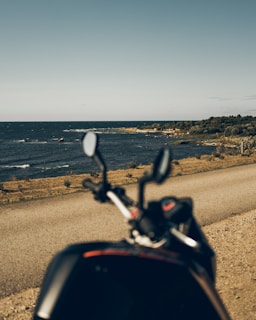 A scenic coastal landscape viewed from a slightly elevated angle, with a road running along the shoreline. The ocean is on the left, meeting a clear sky above. In the foreground, the blurred outline of a motorcycle is visible, suggesting a viewpoint from a biker's perspective.