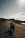 A motorcycle is parked on the side of an empty, winding road under a vast, open sky. The landscape is rugged with patches of vegetation and distant hills, evoking a sense of solitude and adventure.