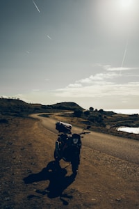 A motorcycle is parked on the side of an empty, winding road under a vast, open sky. The landscape is rugged with patches of vegetation and distant hills, evoking a sense of solitude and adventure.