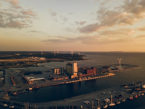 An aerial view of an industrial port area with warehouses and multiple wind turbines. The landscape is bathed in warm sunlight, likely near sunset, creating long shadows. The water around the port reflects some of the orange hues of the sky, and several small boats are moored along the docks.