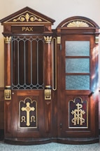 A wooden confessional booth with intricate carvings and religious symbols, including a large cross and the keys of Saint Peter. The word 'PAX' is inscribed at the top. The booth features decorative elements painted in gold, contrasting against the rich brown wood. One side of the booth has a closed door with textured glass panes.