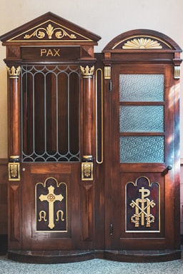 A wooden confessional booth with intricate carvings and religious symbols, including a large cross and the keys of Saint Peter. The word 'PAX' is inscribed at the top. The booth features decorative elements painted in gold, contrasting against the rich brown wood. One side of the booth has a closed door with textured glass panes.