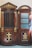 A wooden confessional booth with intricate carvings and religious symbols, including a large cross and the keys of Saint Peter. The word 'PAX' is inscribed at the top. The booth features decorative elements painted in gold, contrasting against the rich brown wood. One side of the booth has a closed door with textured glass panes.