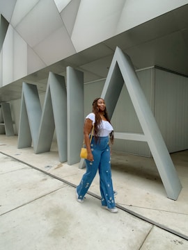 A woman walks past large architectural letters that spell out 'MIA'. She is wearing a white top, blue jeans with decorative designs, sneakers, and carries a yellow handbag. The background features modern architectural elements with angular, metallic surfaces.