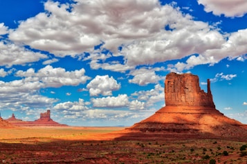 A vast desert landscape featuring a prominent, towering red rock formation under a vibrant blue sky filled with scattered white clouds. The ground is barren with patches of vegetation, and the sunlight casts dramatic shadows on the terrain.