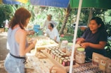 A market stall displays various packaged goods, including jars and bags filled with different products. Two women interact at the stall, with one appearing to make a purchase. Behind the stall, there is a variety of produce and people wearing casual clothing. The setting is outdoors with trees and plants in the background, providing a lush, green environment.