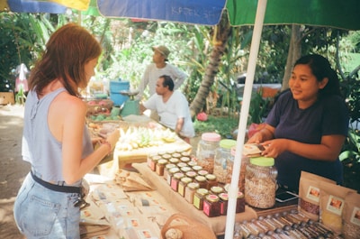 A market stall displays various packaged goods, including jars and bags filled with different products. Two women interact at the stall, with one appearing to make a purchase. Behind the stall, there is a variety of produce and people wearing casual clothing. The setting is outdoors with trees and plants in the background, providing a lush, green environment.