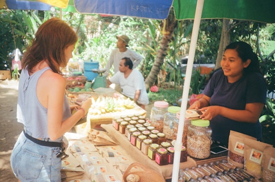 A market stall displays various packaged goods, including jars and bags filled with different products. Two women interact at the stall, with one appearing to make a purchase. Behind the stall, there is a variety of produce and people wearing casual clothing. The setting is outdoors with trees and plants in the background, providing a lush, green environment.