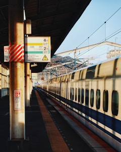 A modern high-speed train at a railway station platform. The image captures the sleek design of the train with a focus on the platform's architecture, including overhead signage and safety notices. The sunlight casts long shadows, suggesting either early morning or late afternoon.