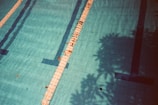 A swimming pool with clear blue water and tiled lanes, featuring lane dividers and some scattered leaves. Shadows of surrounding trees or foliage cast patterns across the water, adding a layer of depth and texture.