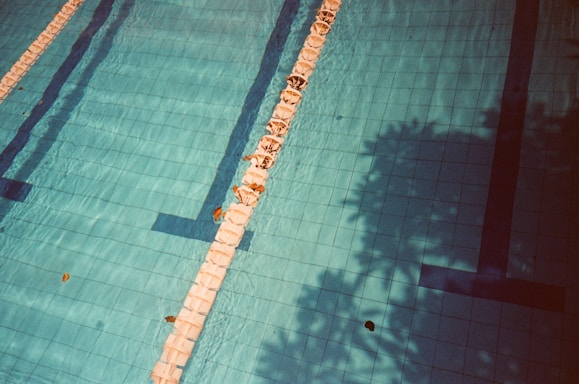 A swimming pool with clear blue water and tiled lanes, featuring lane dividers and some scattered leaves. Shadows of surrounding trees or foliage cast patterns across the water, adding a layer of depth and texture.