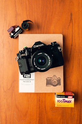 A vintage Canon A-1 film camera is placed on top of a notebook with a sketch of a camera. Nearby, there are film canisters and a box of Kodak T-Max 100 film. The wooden surface provides a warm backdrop.