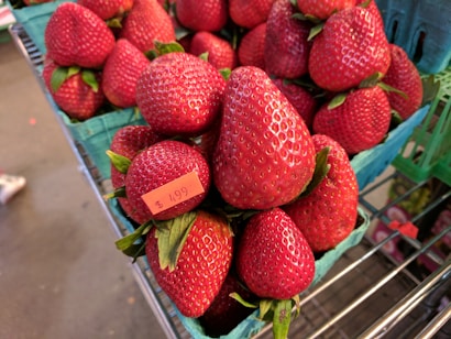Fresh strawberries are displayed in green containers on a metal shelf. A price tag reading $4.99 is visible on one of the containers.