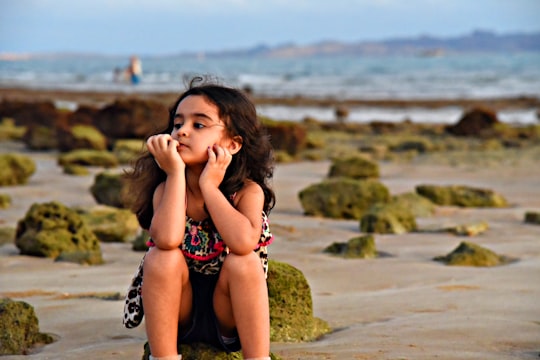 girl wearing black and white with red spaghetti strap top sitting on rock near seashore under blue and white sky in Qeshm Iran