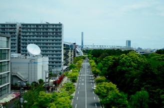 A cityscape featuring a series of high-rise buildings, with one prominently displaying a large satellite dish. An empty road lined with trees runs through the center, leading to a distant bridge in the background. Greenery, including trees and shrubs, is abundant, contrasting with the urban structures.