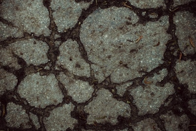 A close-up view of a cracked concrete surface with several irregular fissures. The surface shows scattered dried leaves and small debris, emphasizing the rough and rugged texture of the concrete.