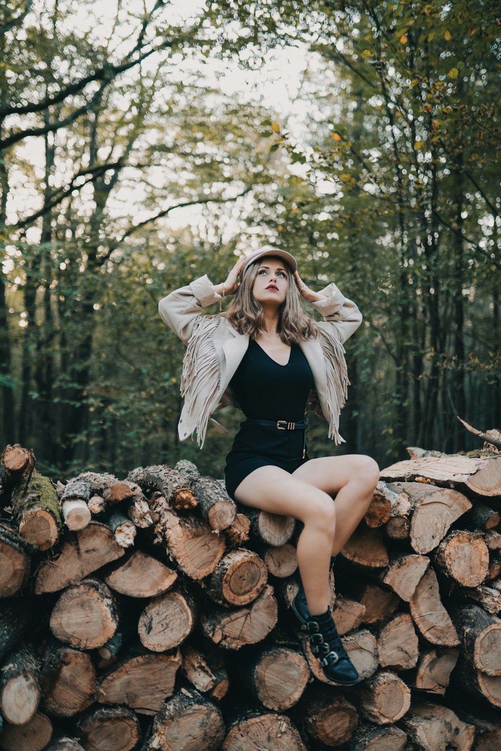 woman sitting on firewood stack looking up while touching her hat surrounded with green trees