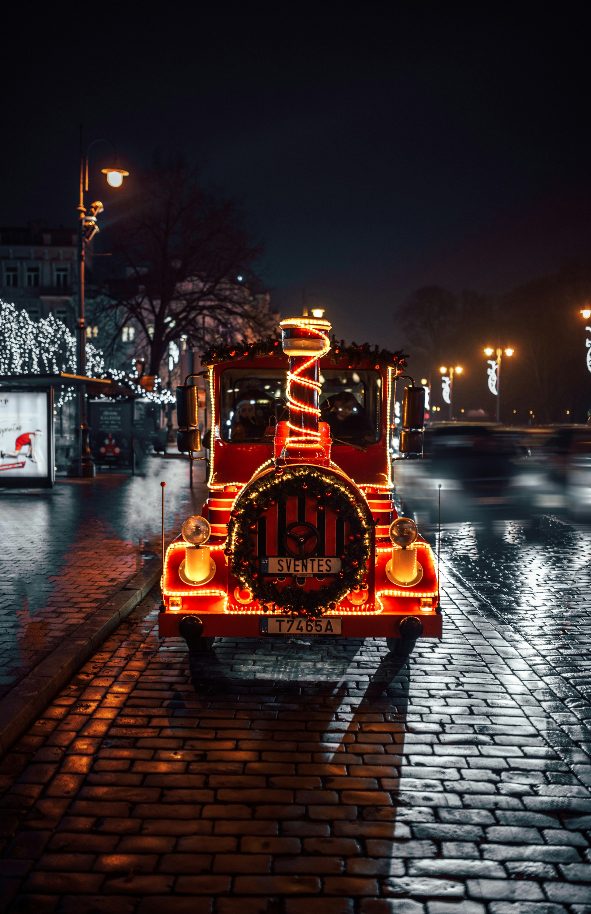 Red festive train decorated with spiraling lights sits on a wet cobblestone street at night, with blurred holiday street decorations in the background.