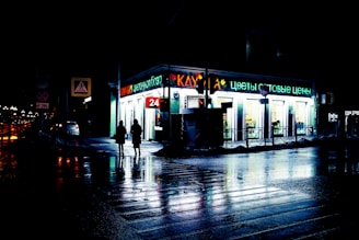 A brightly lit 24-hour store with neon signage in a nighttime urban setting. Two people are silhouetted against the light as they walk by, and the wet pavement reflects the colorful lights, creating a moody atmosphere.