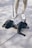 A black French Bulldog is lying down on a concrete surface, wearing a striped backpack. The dog is attached to a leash held by a person wearing beige pants and white sneakers.