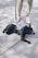 A black French Bulldog is lying down on a concrete surface, wearing a striped backpack. The dog is attached to a leash held by a person wearing beige pants and white sneakers.