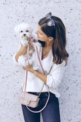 A woman in a white blouse holds a small white dog in her arms. She is wearing dark jeans and sunglasses on her head. A light pink purse with a gold chain is slung across her shoulder. The background is a speckled light gray wall.
