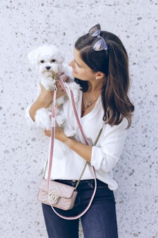A woman in a white blouse holds a small white dog in her arms. She is wearing dark jeans and sunglasses on her head. A light pink purse with a gold chain is slung across her shoulder. The background is a speckled light gray wall.