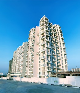 A tall residential apartment building stands prominently against a clear blue sky. The structure features multiple balconies and is painted in a light cream color. The foreground shows a spacious, empty pavement and a low boundary wall.