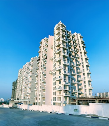 A tall residential apartment building stands prominently against a clear blue sky. The structure features multiple balconies and is painted in a light cream color. The foreground shows a spacious, empty pavement and a low boundary wall.