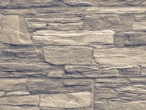 An arrangement of rectangular stones laid out in layers, forming a textured wall pattern. The stones vary in size and exhibit a rugged, rough appearance with natural lines and grooves.