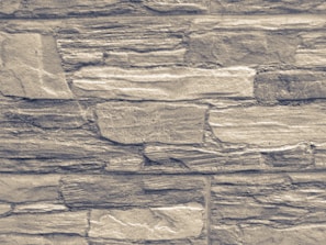 An arrangement of rectangular stones laid out in layers, forming a textured wall pattern. The stones vary in size and exhibit a rugged, rough appearance with natural lines and grooves.