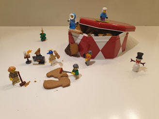 Several Lego figures are depicted around an open red and white tin filled with cookies. Some figures appear to be interacting with the cookies, with one holding an axe and another holding a mug. Additional elements include a snowman figure, a miniature Christmas tree, and a small fireplace setup. The scene conveys a playful and imaginative atmosphere.