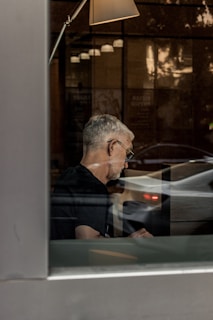A person sits indoors, visible through a window, focused on an unseen task. The setting is illuminated by the soft glow of a lamp, and there are reflections of outside traffic creating a juxtaposition of indoor calm and outdoor motion.