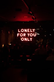A dimly lit room featuring a neon red sign that reads 'LONELY FOR YOU ONLY.' The ambiance is dark, with subtle lighting highlighting the silhouettes of people sitting at tables, creating an intimate and mysterious atmosphere.