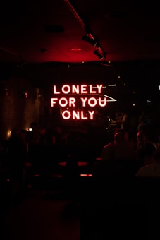 A dimly lit room featuring a neon red sign that reads 'LONELY FOR YOU ONLY.' The ambiance is dark, with subtle lighting highlighting the silhouettes of people sitting at tables, creating an intimate and mysterious atmosphere.