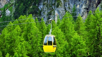 A bright yellow cable car glides along a cable against a backdrop of lush green forest. Rocky cliffs can be seen in the distance, adding a rugged element to the serene natural scene.