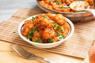 A bowl filled with crispy, golden-brown fried dish covered in a rich, spicy sauce, topped with green herbs. The background includes a bamboo mat and a fork, with a portion of the dish also visible in a pan.