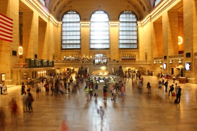 A grand hall with high arched windows letting in natural light, bustling with people moving in various directions. The flooring is marble, and the walls are adorned with ornate details. A large flag is prominently displayed to the left, and a circular information booth is centrally located.