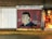 A mural on a brick wall featuring a portrait of a person with a cross and the name Emilio Falcon next to it. The years 1977 and 2018 are written on the mural. A bicycle is parked in front of the mural. To the right, there is an entrance leading into a brightly lit shop.