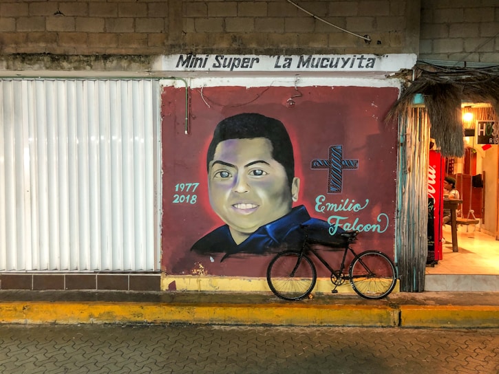A mural on a brick wall featuring a portrait of a person with a cross and the name Emilio Falcon next to it. The years 1977 and 2018 are written on the mural. A bicycle is parked in front of the mural. To the right, there is an entrance leading into a brightly lit shop.