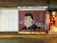 A mural on a brick wall featuring a portrait of a person with a cross and the name Emilio Falcon next to it. The years 1977 and 2018 are written on the mural. A bicycle is parked in front of the mural. To the right, there is an entrance leading into a brightly lit shop.