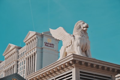 A large building with classical architecture and columns is topped by a statue of a winged lion. The building has an emblem and the words 'Caesars Palace' displayed prominently. The sky is clear and blue, providing a backdrop to the ornate structure.